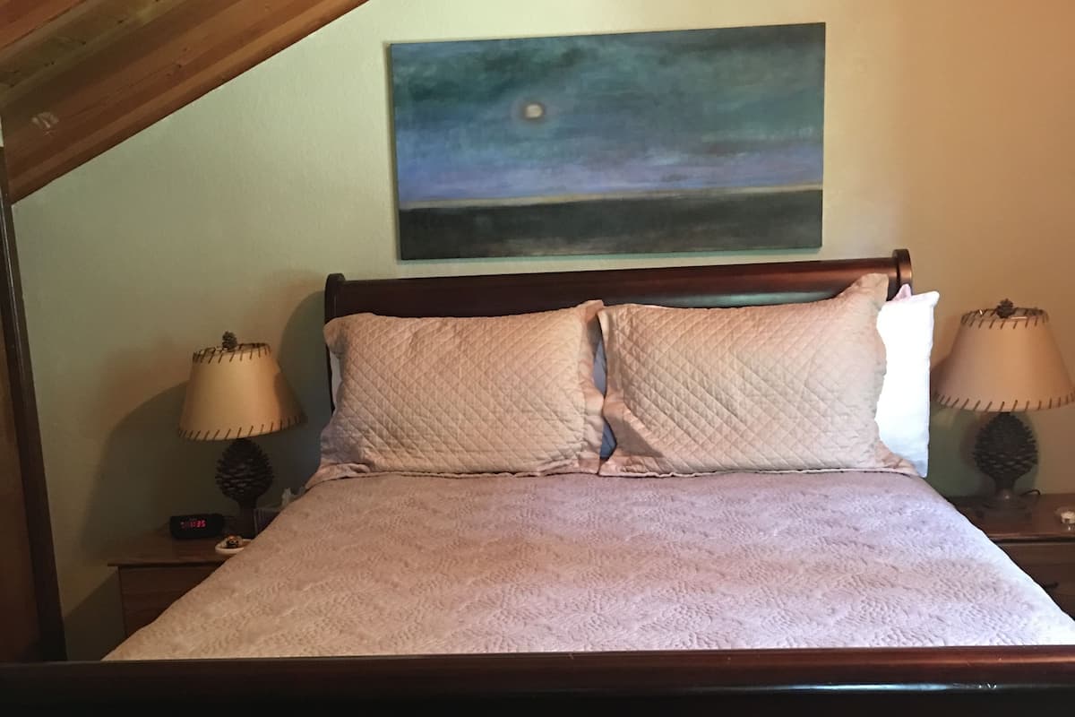 A bedroom with a wooden ceiling, a bed with a quilted mattress, two bedside lamps, and a painting on the wall.
