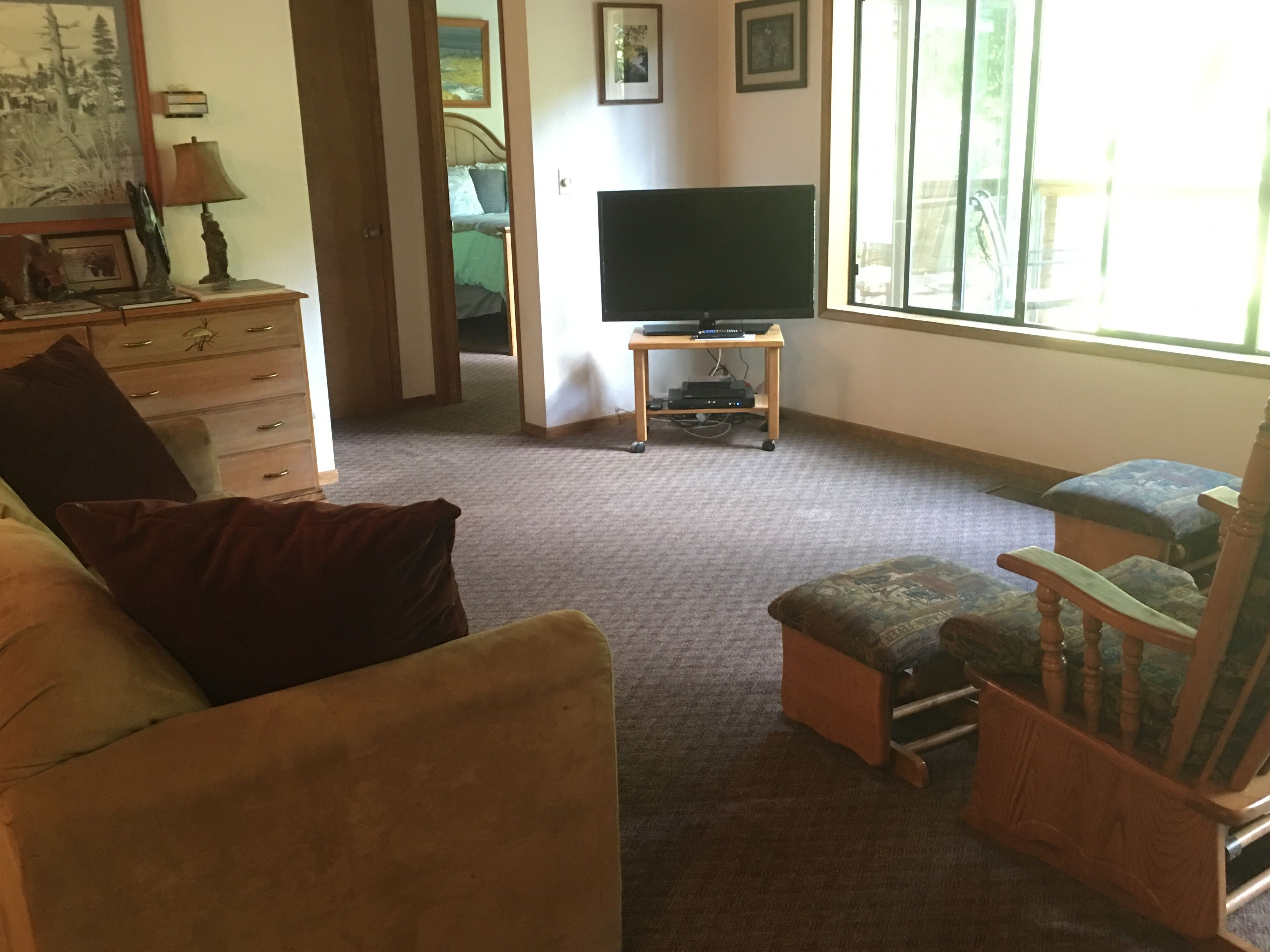 A living room with a sofa, ottoman, wooden chair, a television on a stand, a dresser, and a painting on the wall.