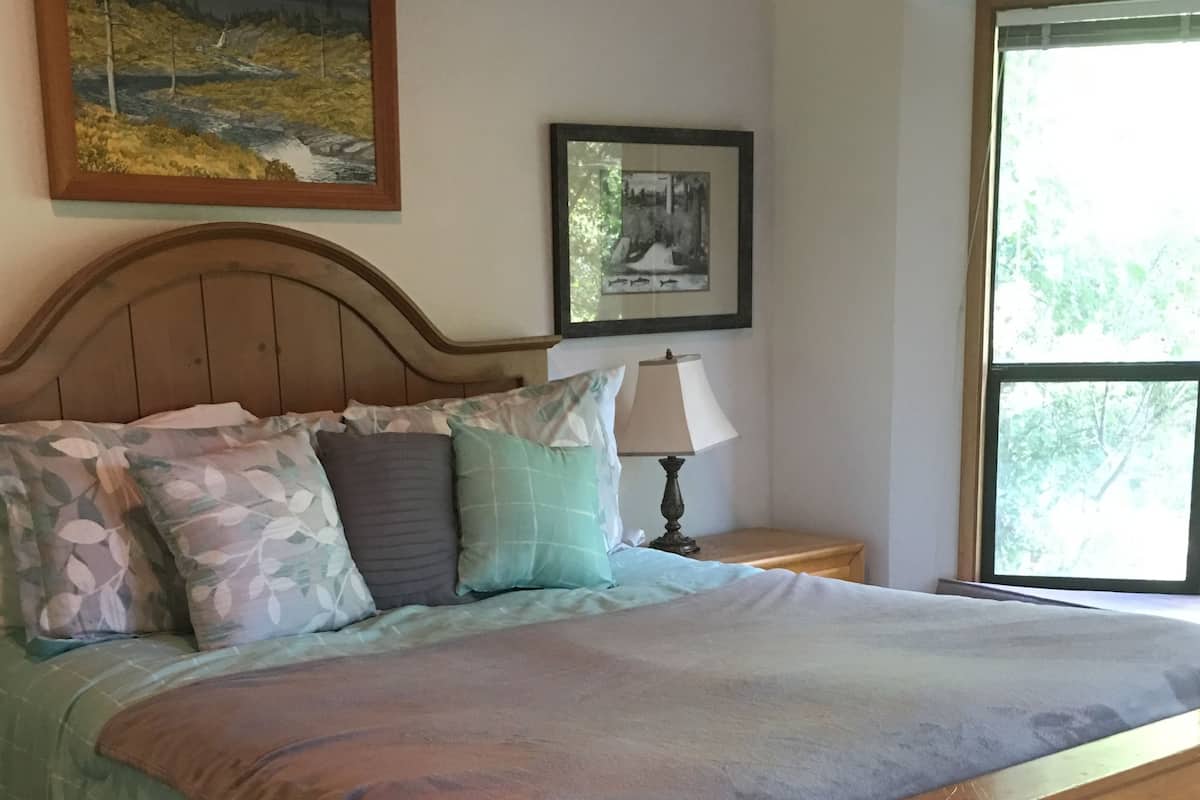A bedroom with a wooden bed, a nightstand with a lamp, a window with a view of trees, and two framed pictures on the wall.