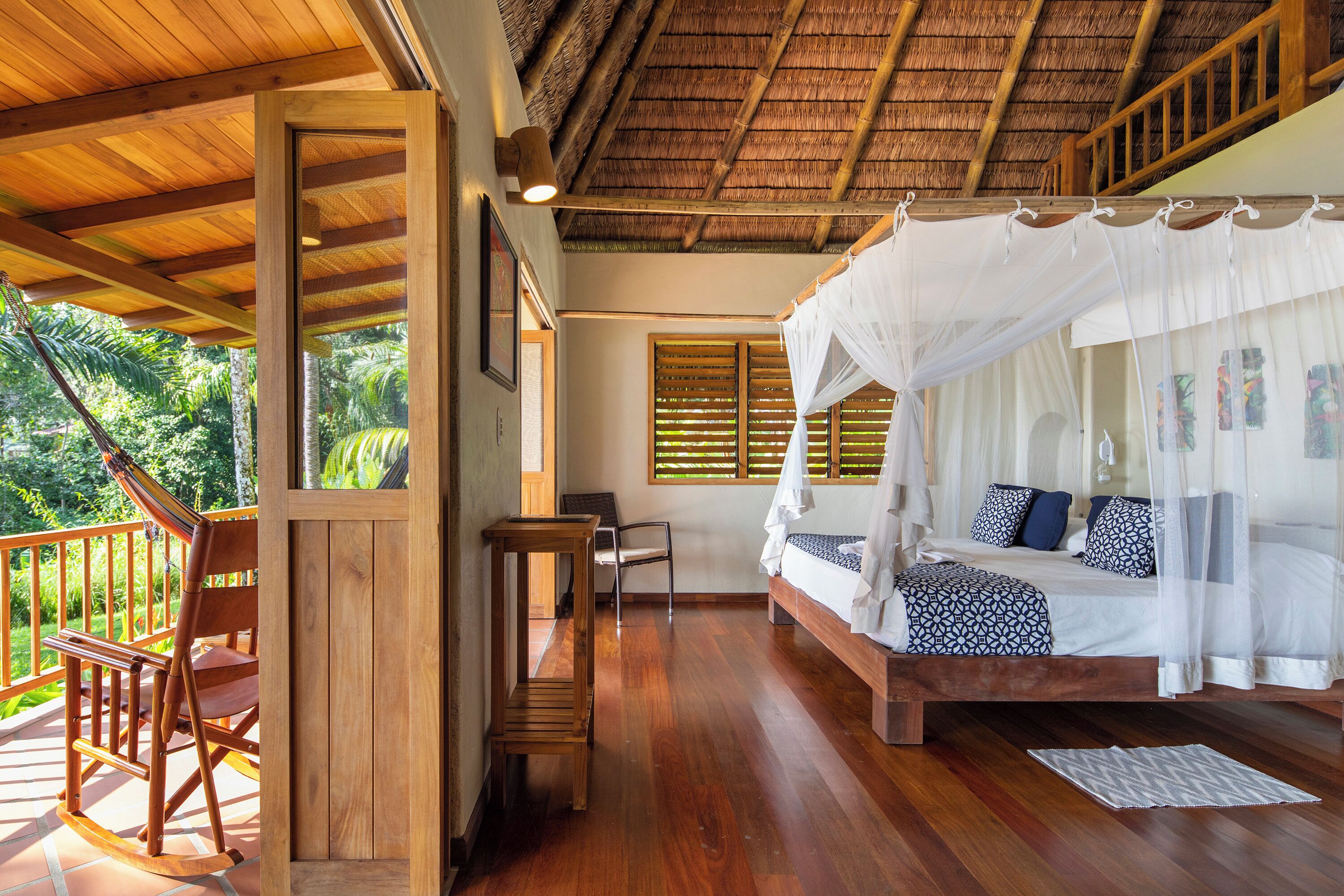 A wooden-framed room with a bed under a canopy, a rocking chair, and a view of lush greenery outside.