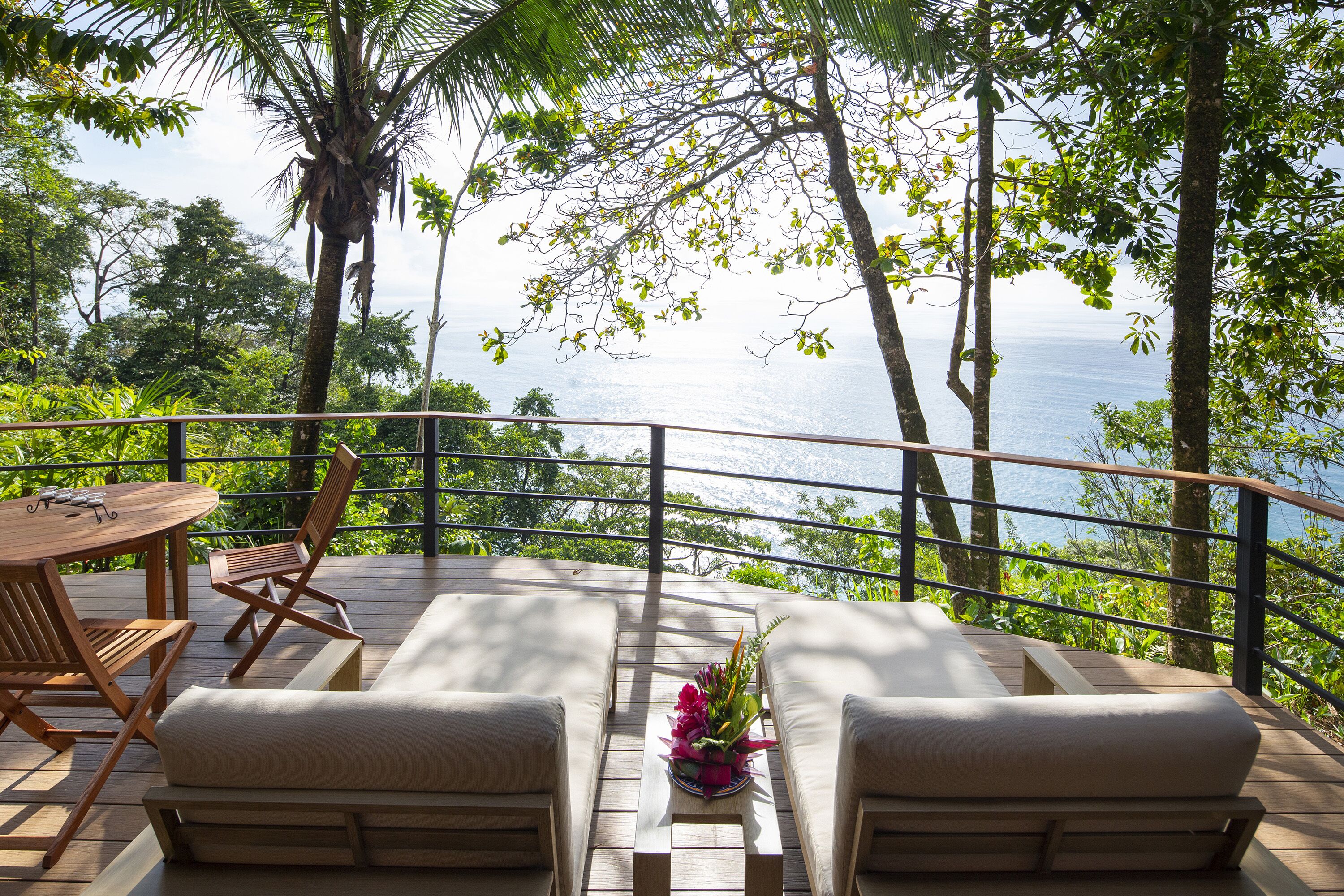 A wooden deck with a table and chairs, overlooking a scenic view of the ocean and lush greenery.