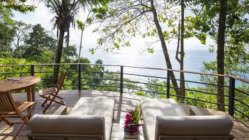 A wooden deck with a table and chairs, overlooking a scenic view of the ocean and lush greenery.