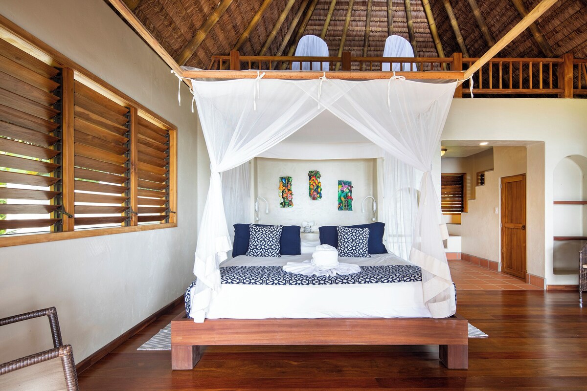 A bed with a canopy, wooden floor, and a view of a room with a door and a chair.