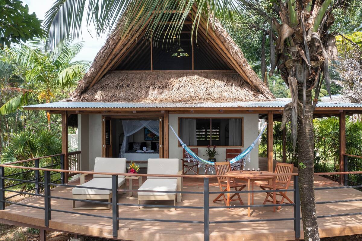 A thatched-roof bungalow with a hammock, outdoor seating, and lush tropical surroundings.