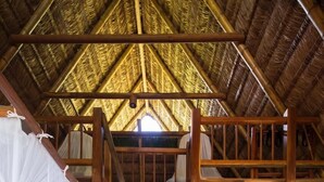 A room with a thatched roof, a wooden staircase, a bed with a mosquito net, a bench, and a plant.