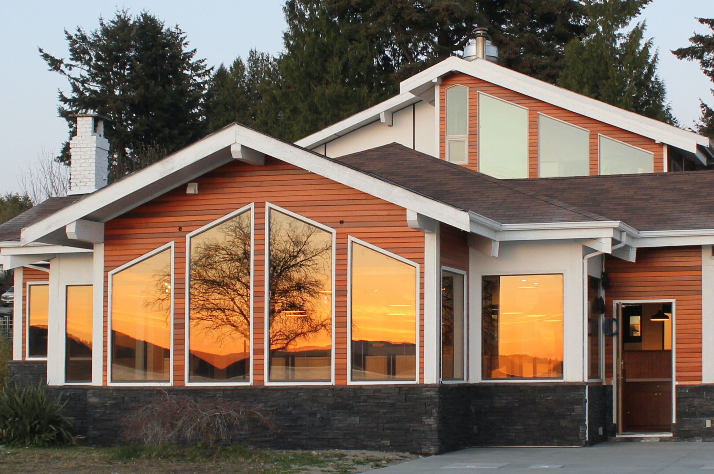 Ein modernes, zweistöckiges Haus mit großen Fenstern, die einen Sonnenuntergang reflektieren, einem Steinfundament und einer Holzfassade.