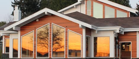A modern two-story house with large windows reflecting a sunset, a stone foundation, and a wooden exterior.