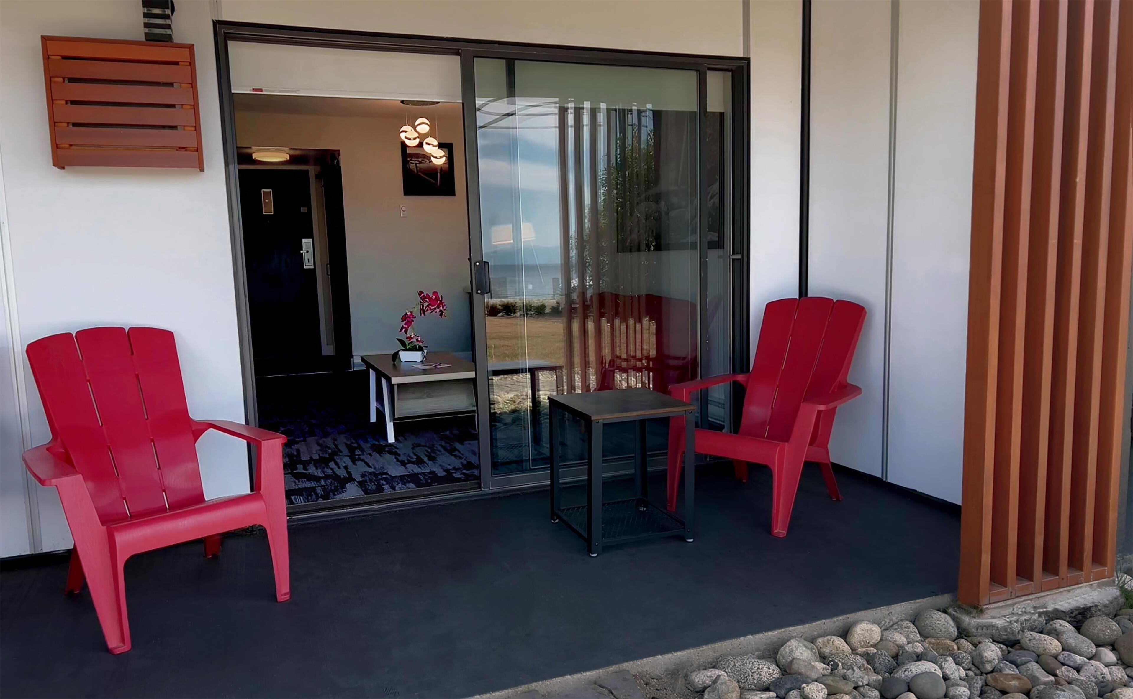 Een balkon met twee rode stoelen, een kleine tafel en een glazen deur die leidt naar een binnenruimte met een plant en een lamp.
