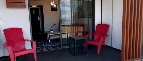 Een balkon met twee rode stoelen, een kleine tafel en een glazen deur die leidt naar een binnenruimte met een plant en een lamp.