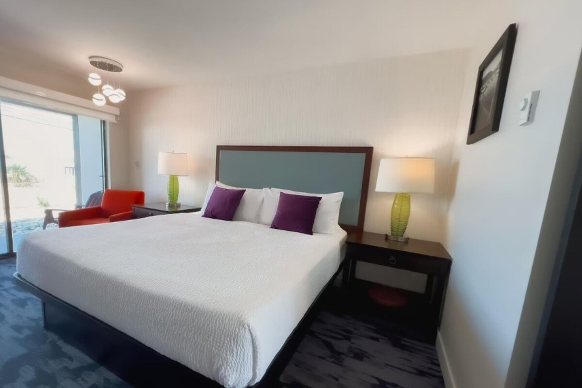 A hotel room with a large bed, two bedside tables with lamps, a red chair, and a view through a large window.