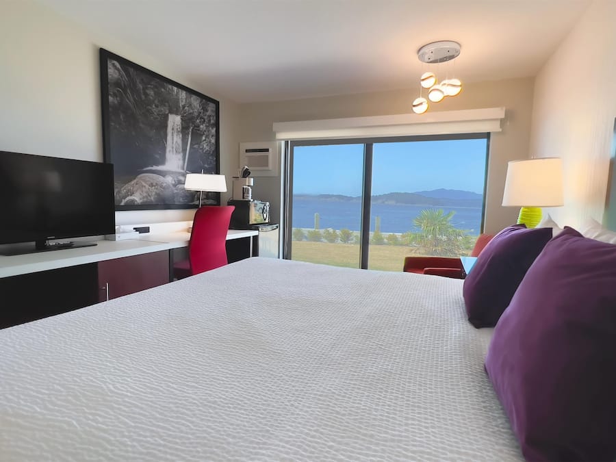 Habitación de hotel con una cama grande, televisión, escritorio con computadora, una silla roja y vista al mar.