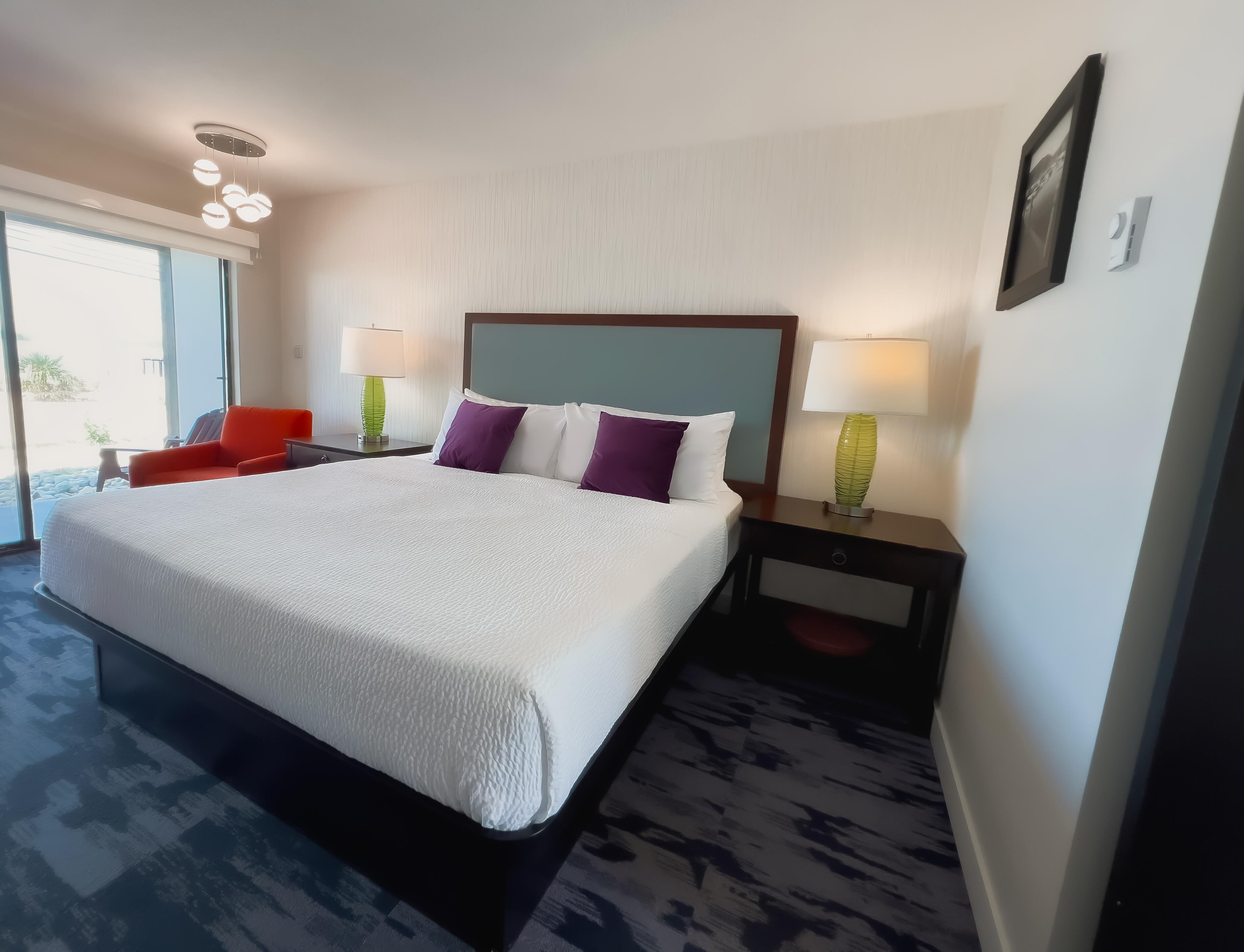 A hotel room with a large bed, two nightstands with lamps, a red chair, and a view through the window.