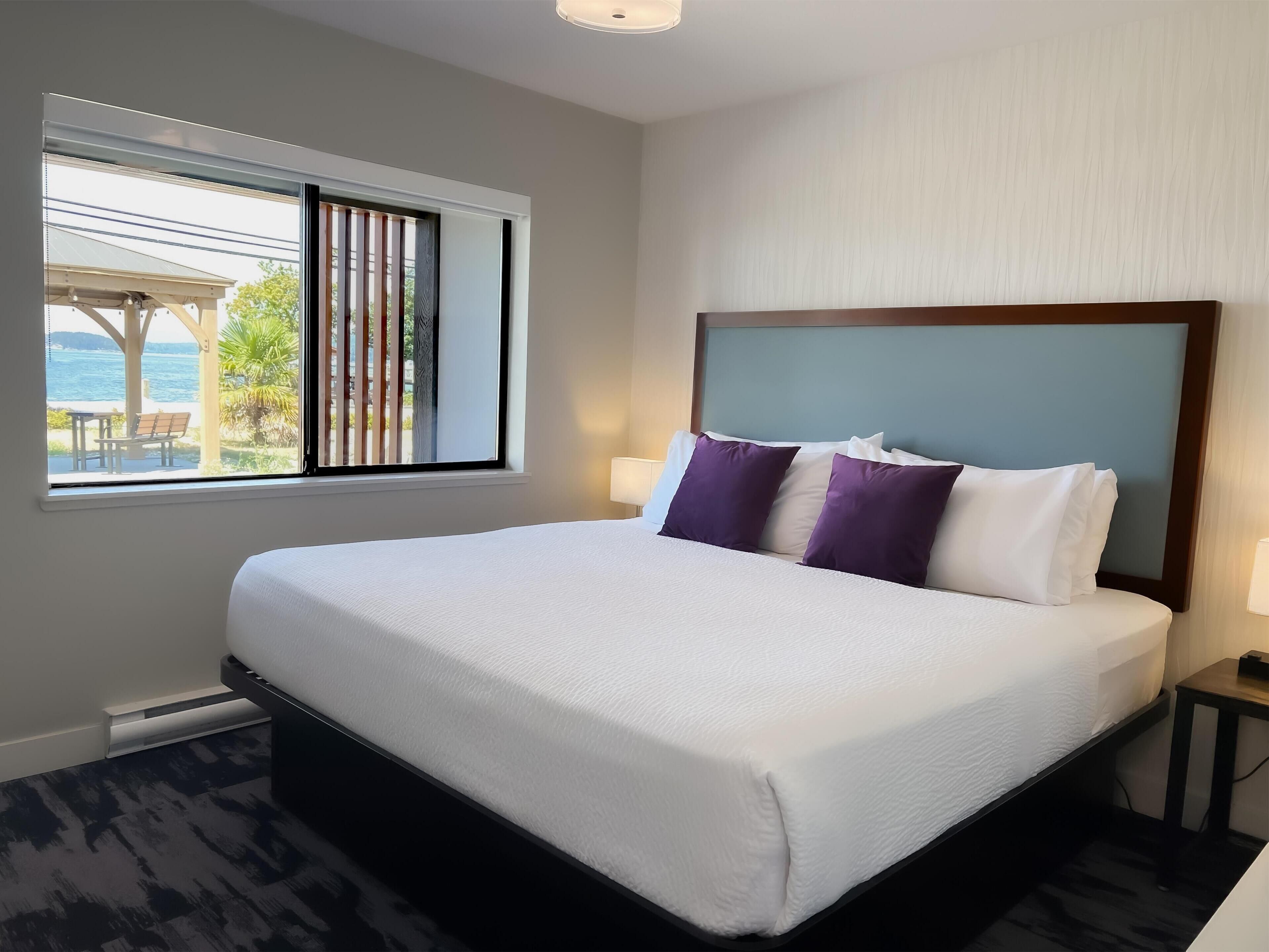 A neatly made bed with white and purple pillows, a wooden headboard, and a view of the sea through a window.
