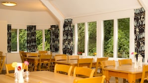 A dining area with wooden tables and chairs, large windows with curtains, and a view of greenery outside. - YHA Grasmere Butharlyp Howe - Hostel (Ambleside)
