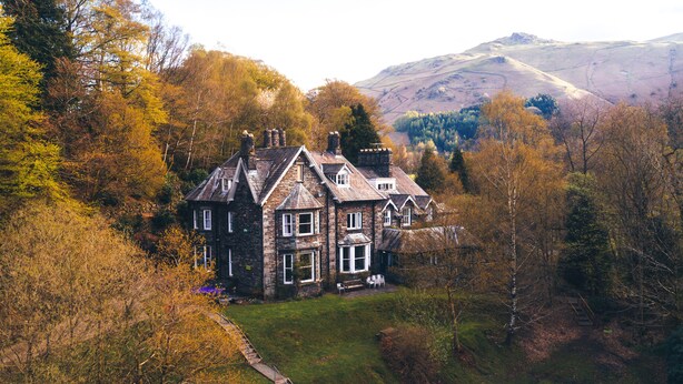 A large, historic stone house with multiple chimneys, surrounded by autumn foliage and a mountainous backdrop. - YHA Grasmere Butharlyp Howe - Hostel (Ambleside)