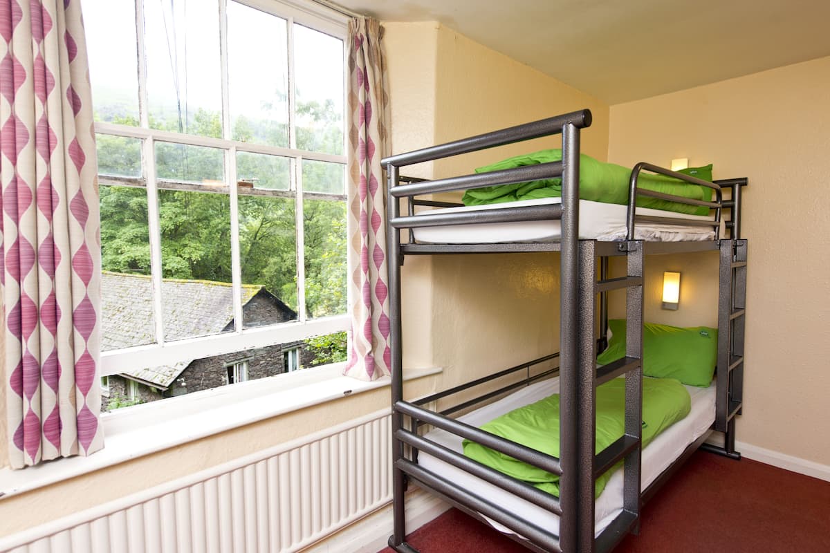 A bunk bed with green bedding, a window with patterned curtains, and a view of greenery outside.