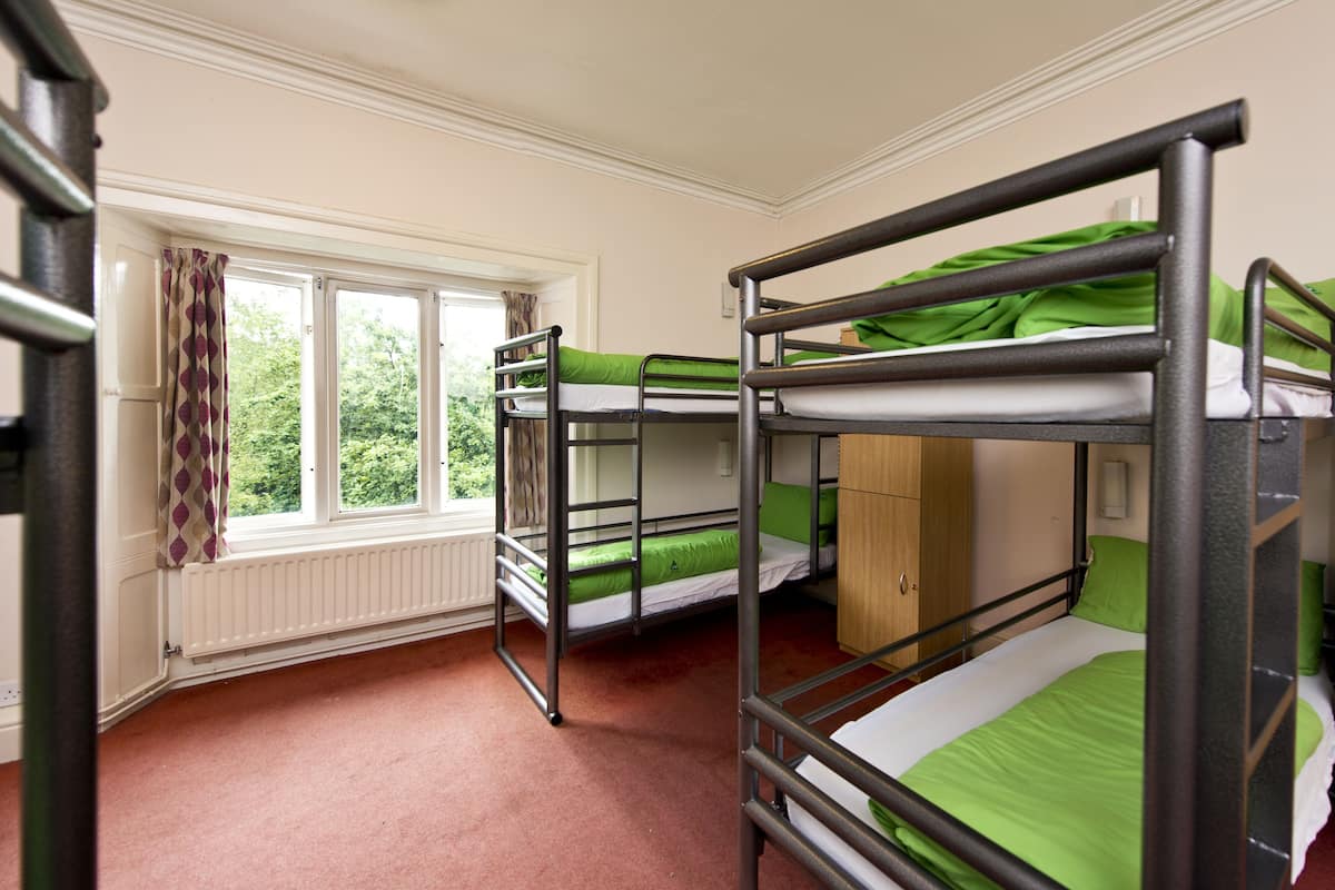 A dormitory room with bunk beds, a window with curtains, a radiator, and a wooden cabinet.