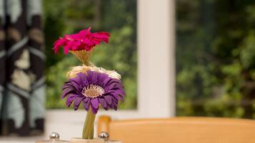 A white vase with two flowers on a wooden table, with salt and pepper shakers and a window in the background.