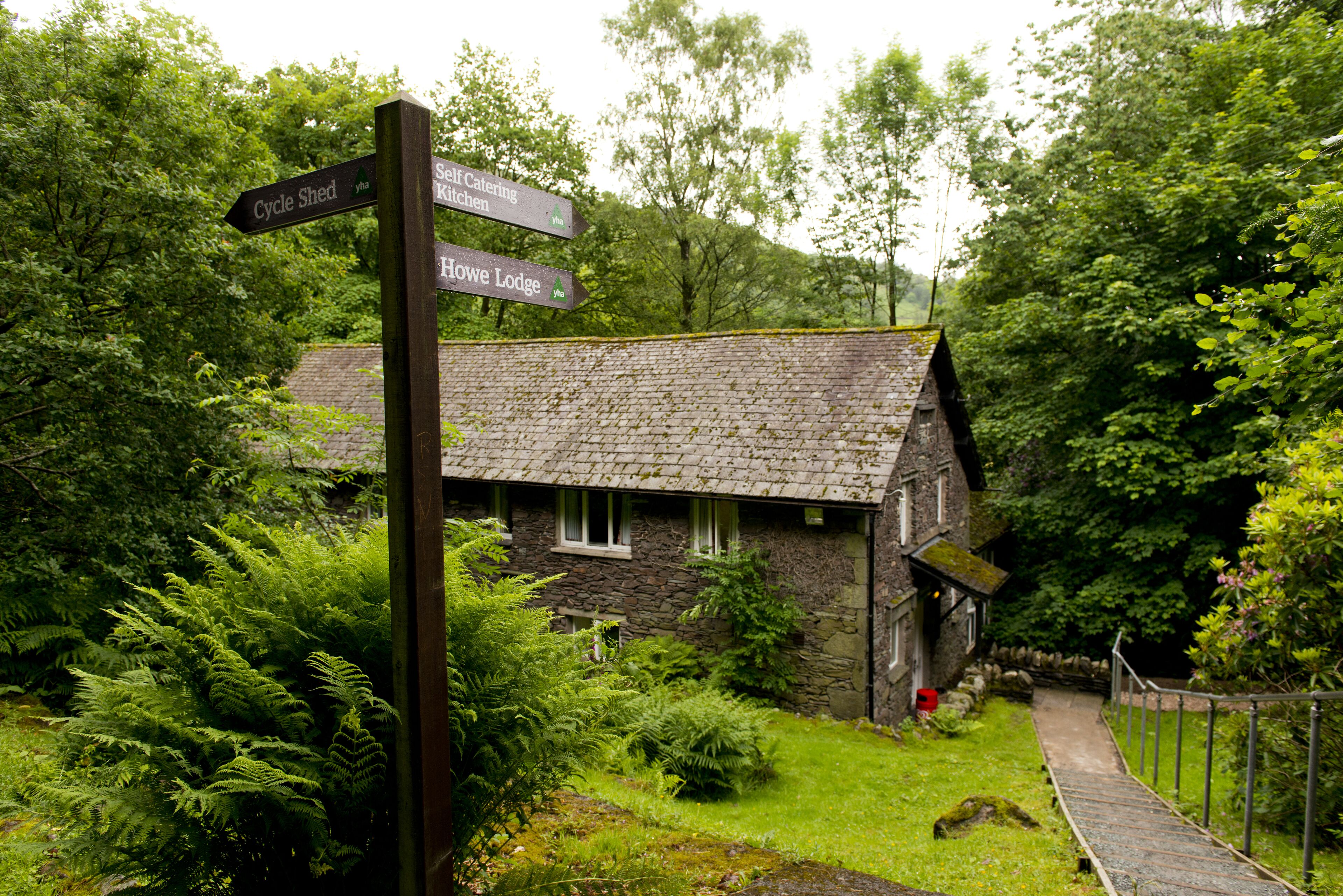 A stone cottage with a thatched roof, surrounded by lush greenery and ferns. A wooden signpost indicates directions to 'Cycle Shed', 'Self Catering Kitchen', and 'Howe Lodge'.