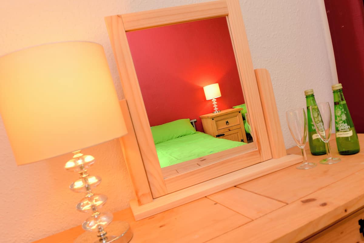 A wooden bedside table with a lamp, a mirror reflecting a room with a bed, and two bottles of green beverage.