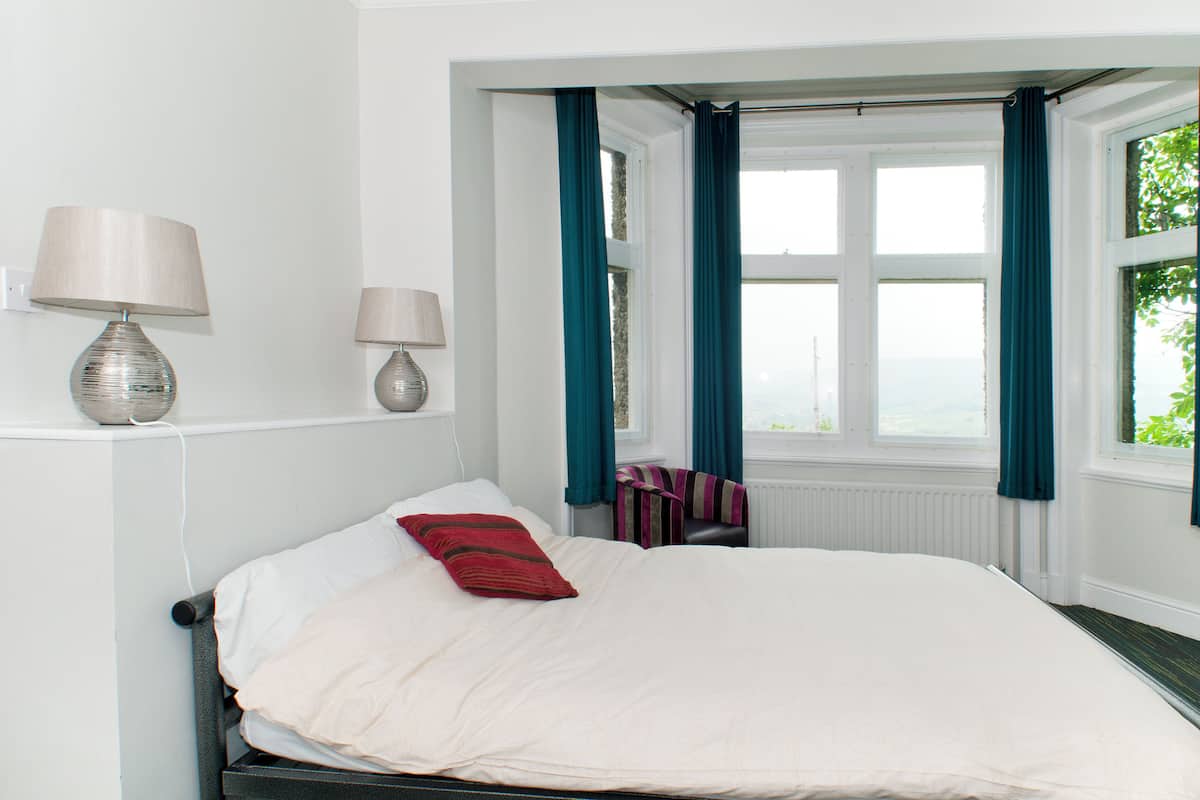 A neatly made bed with a red pillow, two bedside lamps, and a window with teal curtains.