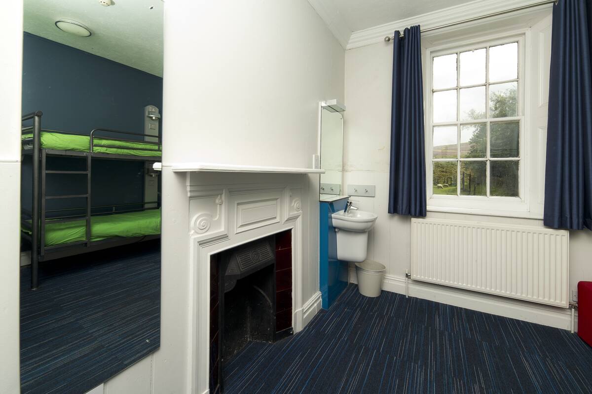 A dormitory room with bunk beds, a fireplace, a sink, and a window with curtains.