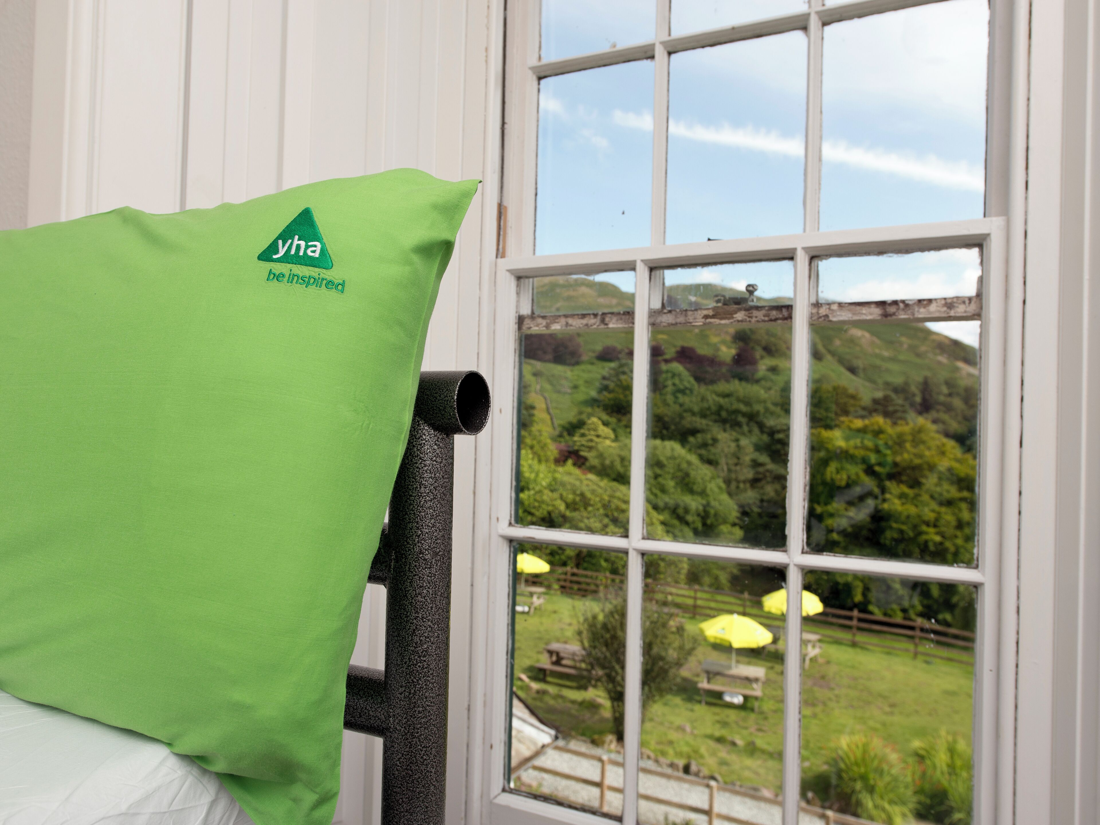 Un almohadón verde con un logotipo y texto, una ventana que da a un paisaje verde, y una pared blanca.