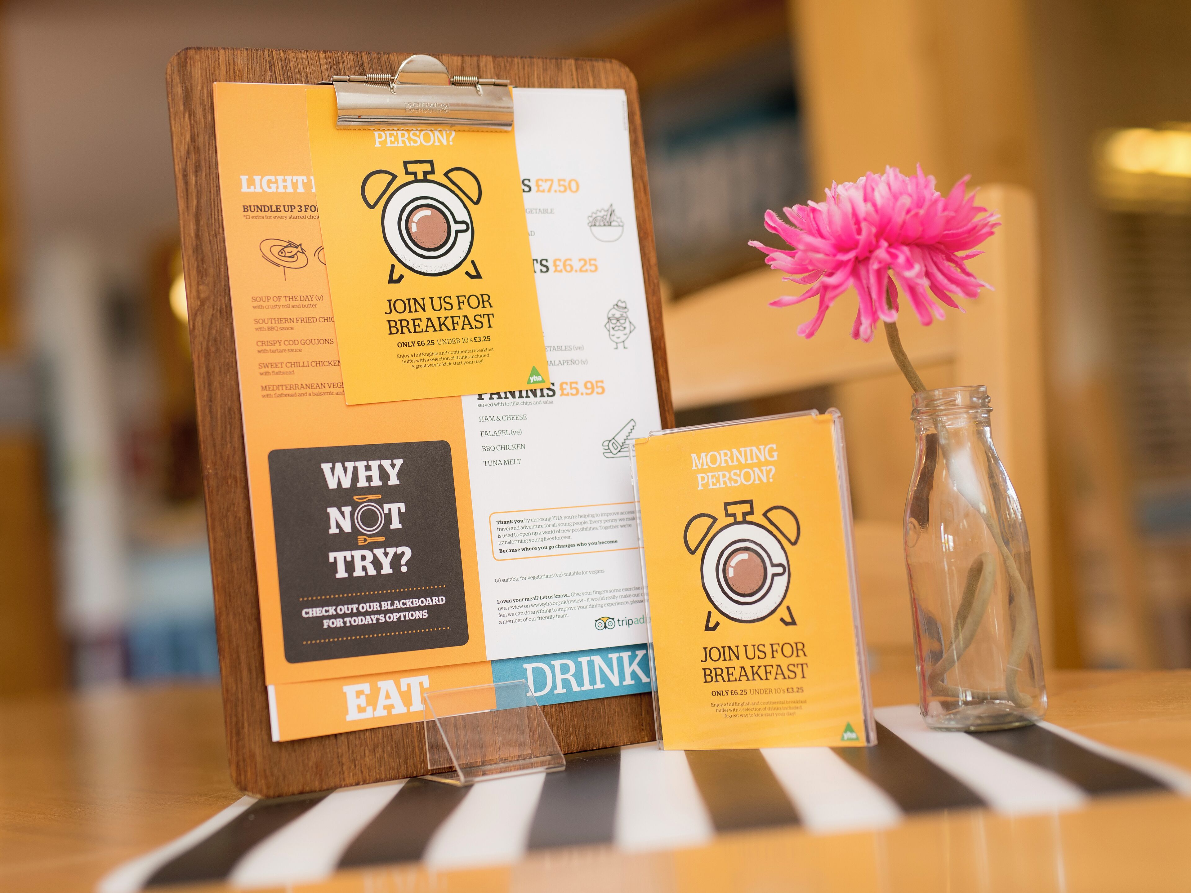 A breakfast menu board with options like 'Light', 'Mediterranean', and 'Why Not Try?' along with a 'Morning Person?' sign and a pink flower in a glass vase.