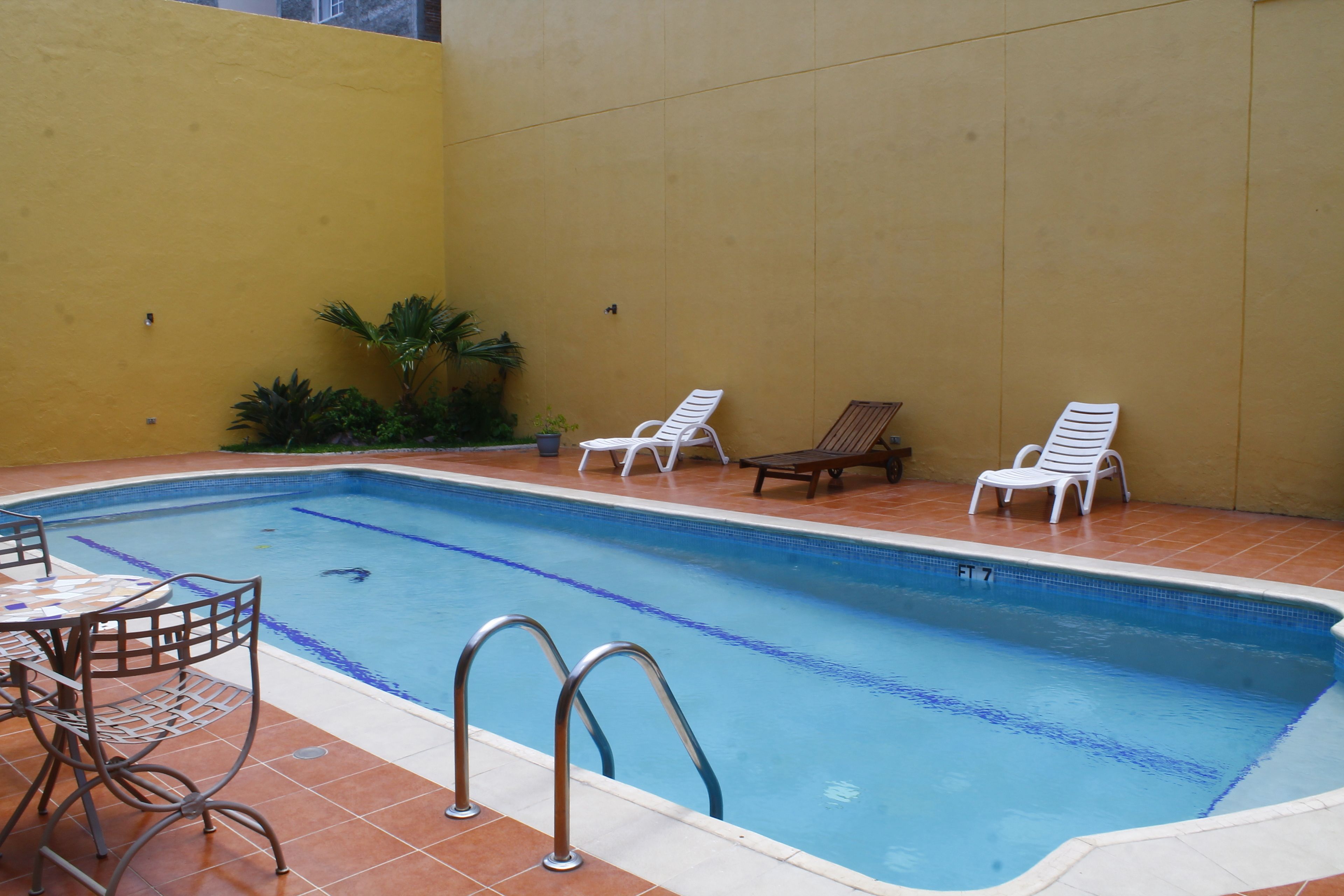 A rectangular indoor swimming pool with a tiled edge, surrounded by a yellow wall and a tiled floor. There are two white lounge chairs and a small table with chairs.
