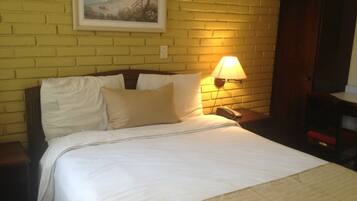 Un lit bien fait, avec du linge de lit blanc, un oreiller beige et une tête de lit en bois. Une lampe fixée au mur assure l’éclairage de la table de chevet.