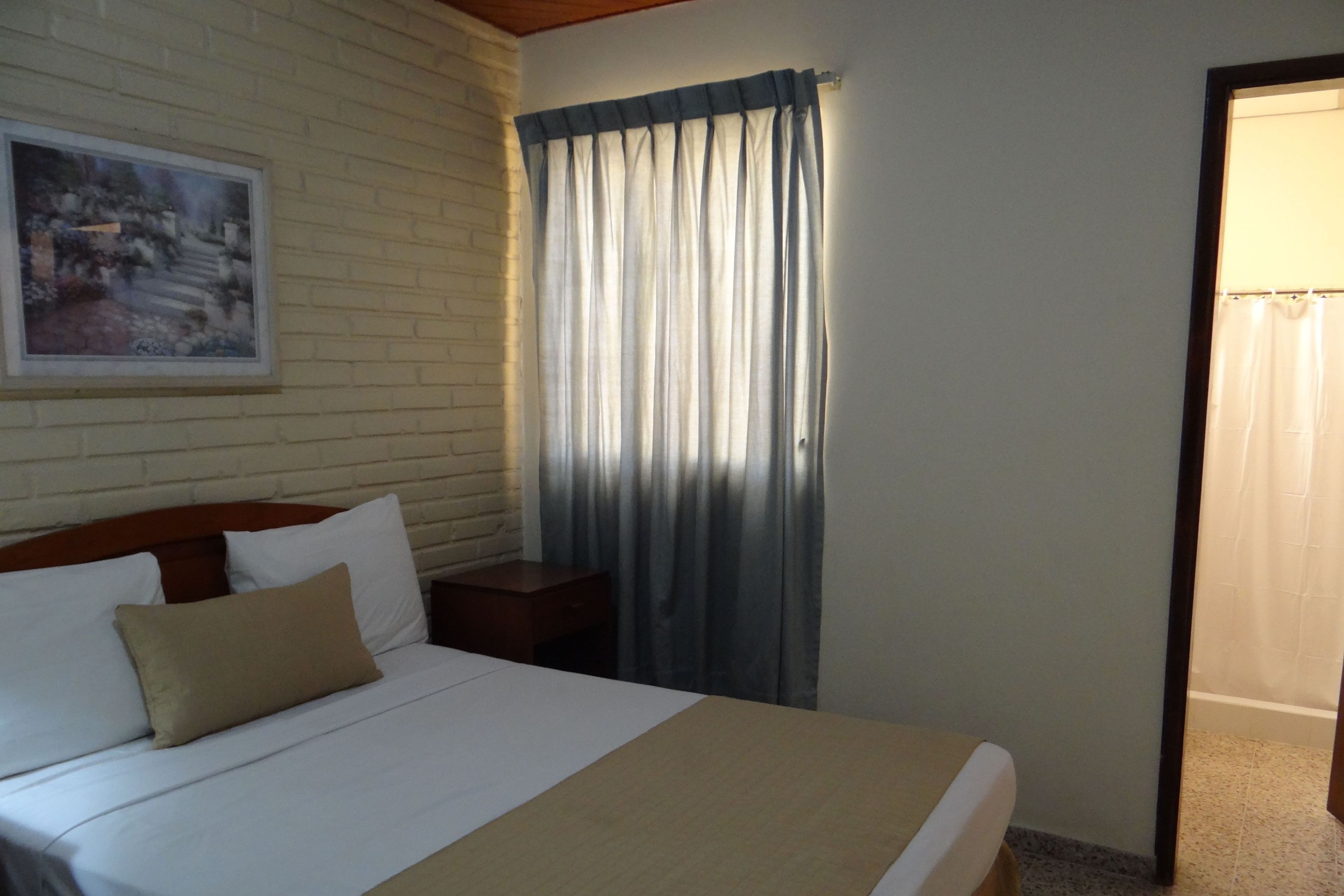 A neatly made bed with white and beige bedding, a wooden headboard, a nightstand with a lamp, a painting on the wall, and a window with curtains.
