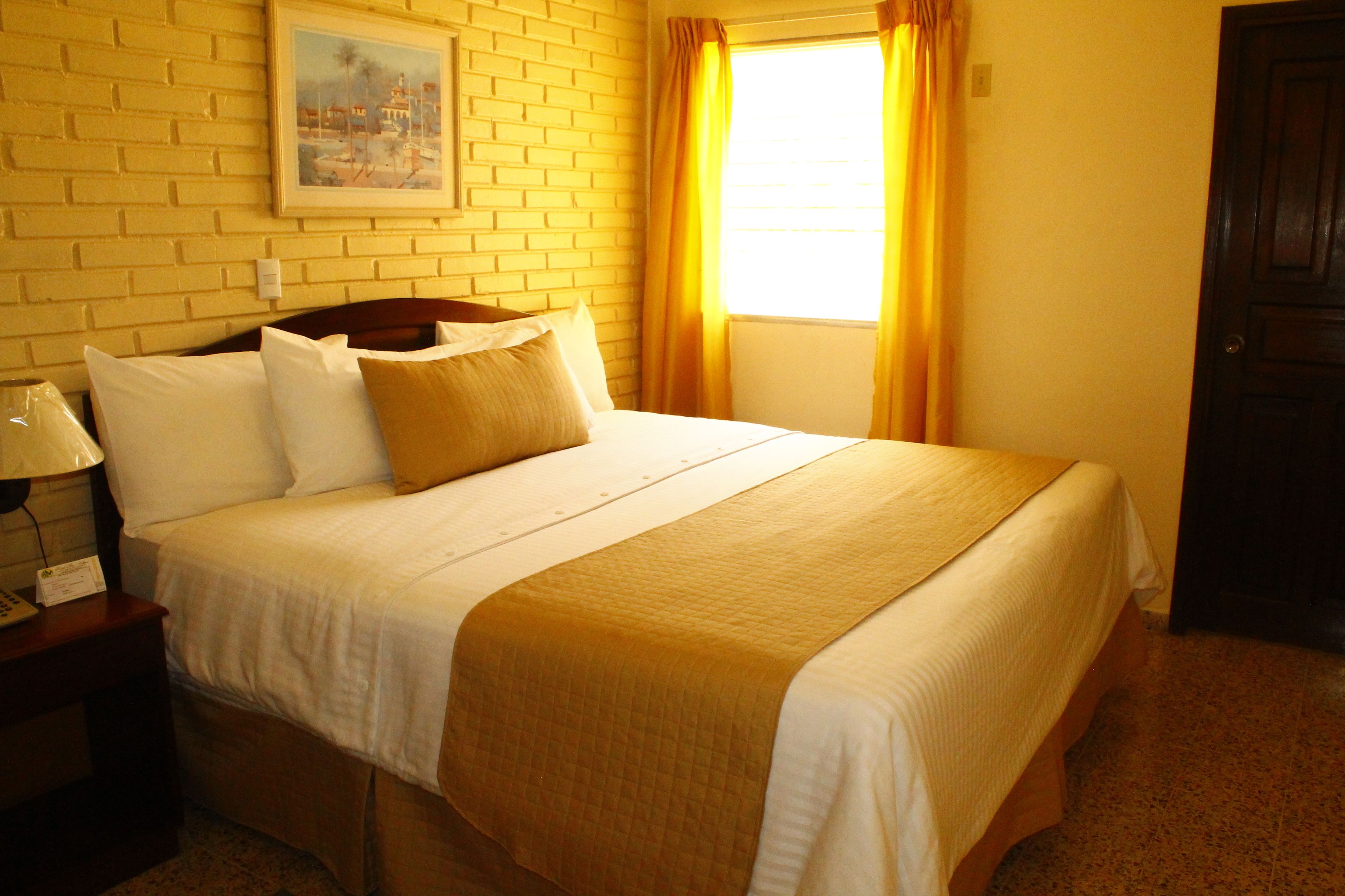 Habitación de hotel con pared de ladrillo, cama con ropa de cama blanca y marrón, mesita de noche con lámpara y ventana con cortinas.