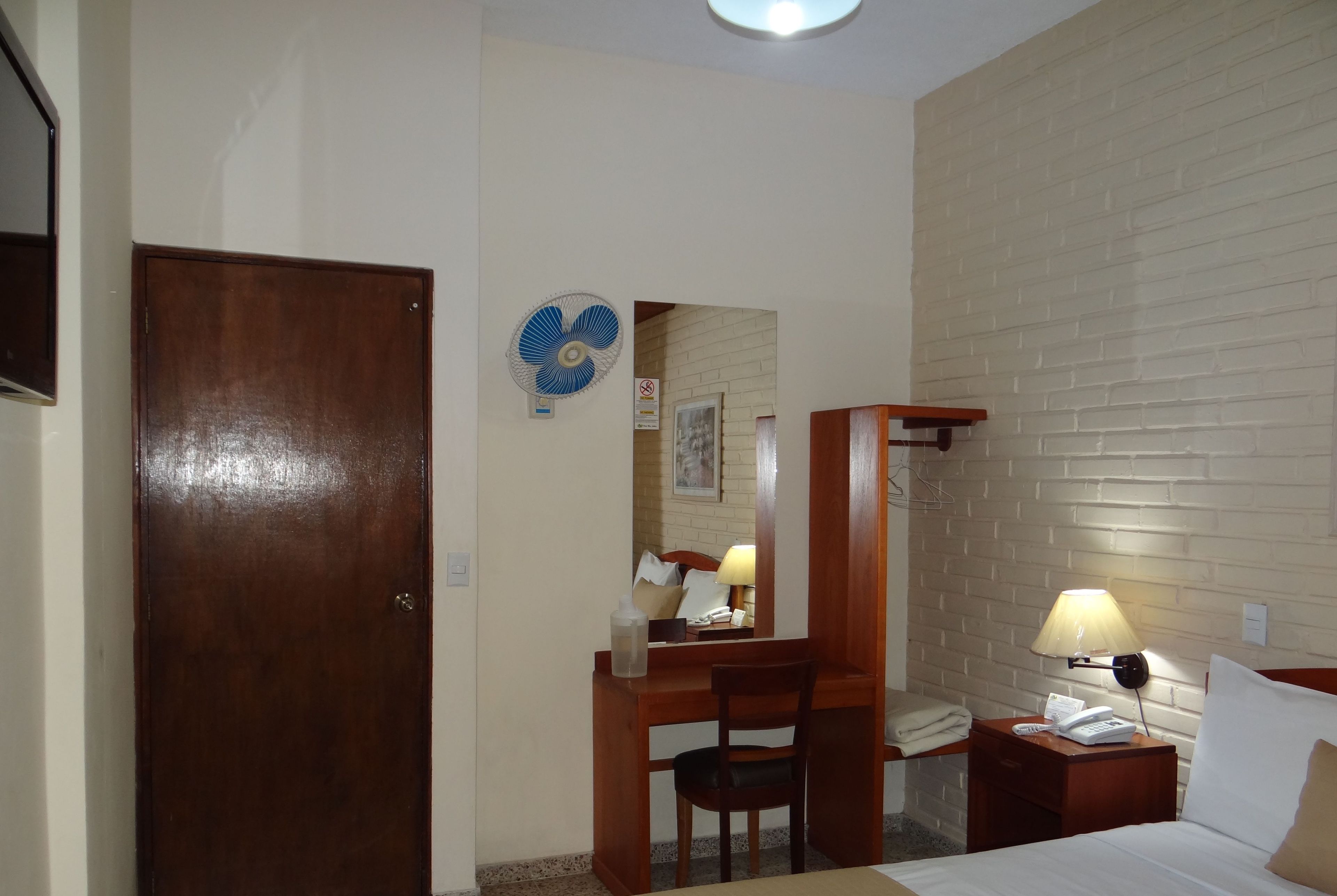 A hotel room with a wooden desk, chair, bedside table with a lamp, a bed, a wall-mounted fan, and a brick-patterned wall.