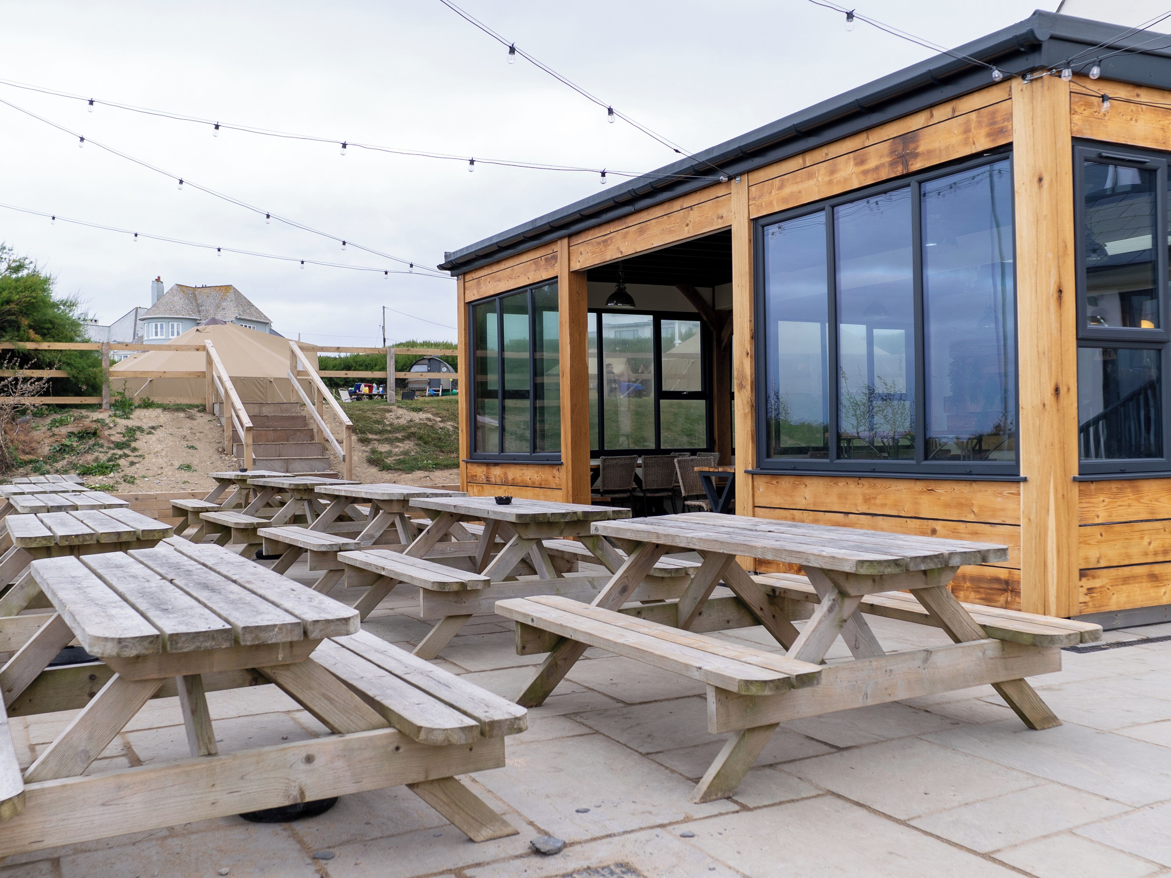An outdoor wooden building with large windows, picnic tables, and a staircase leading to a higher level.