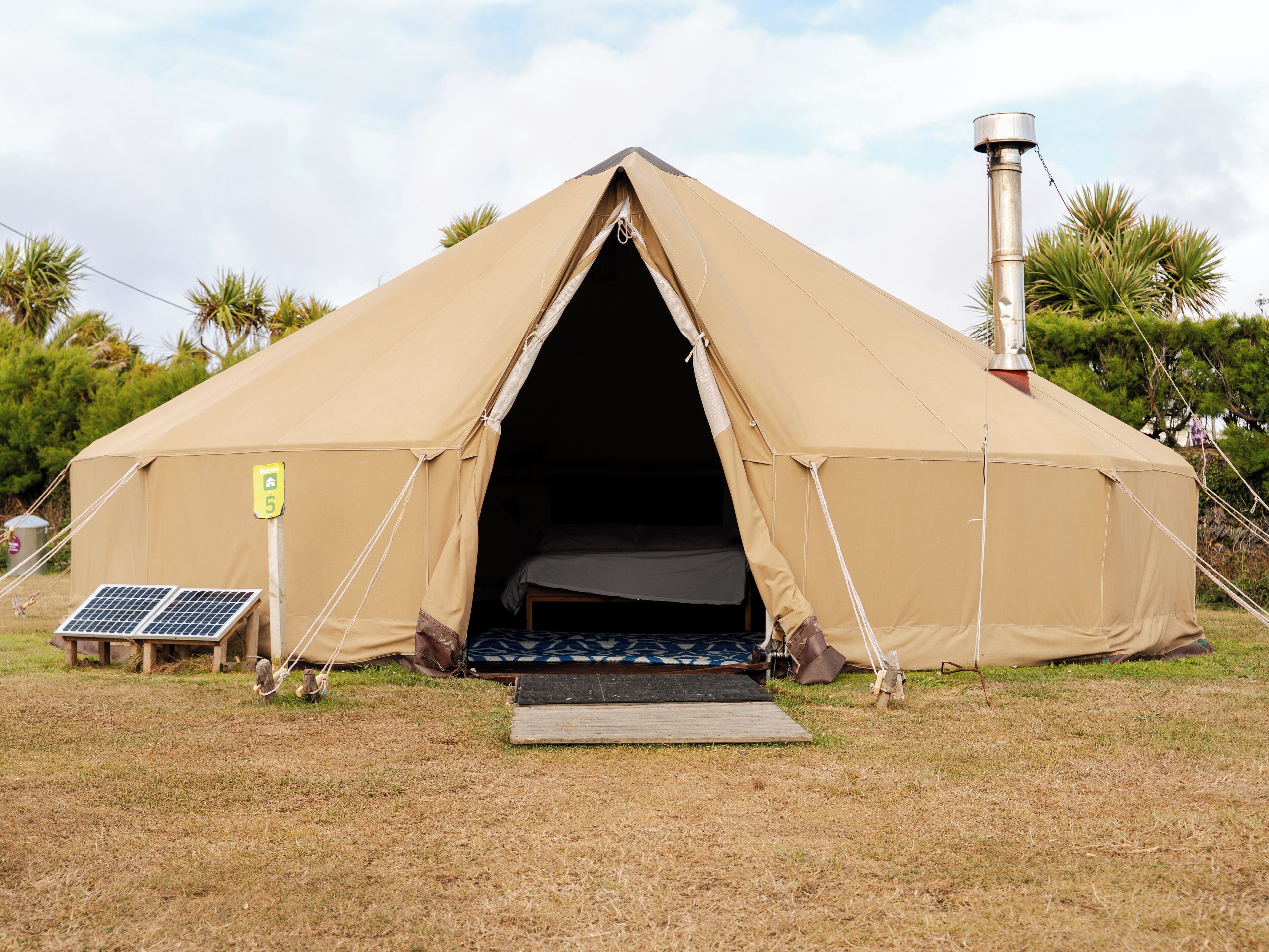 Een grote, beige tent met een deur en een kleine hellingbaan die erheen leidt. Er ligt een zonnepaneel en een metalen schoorsteen op de grond.