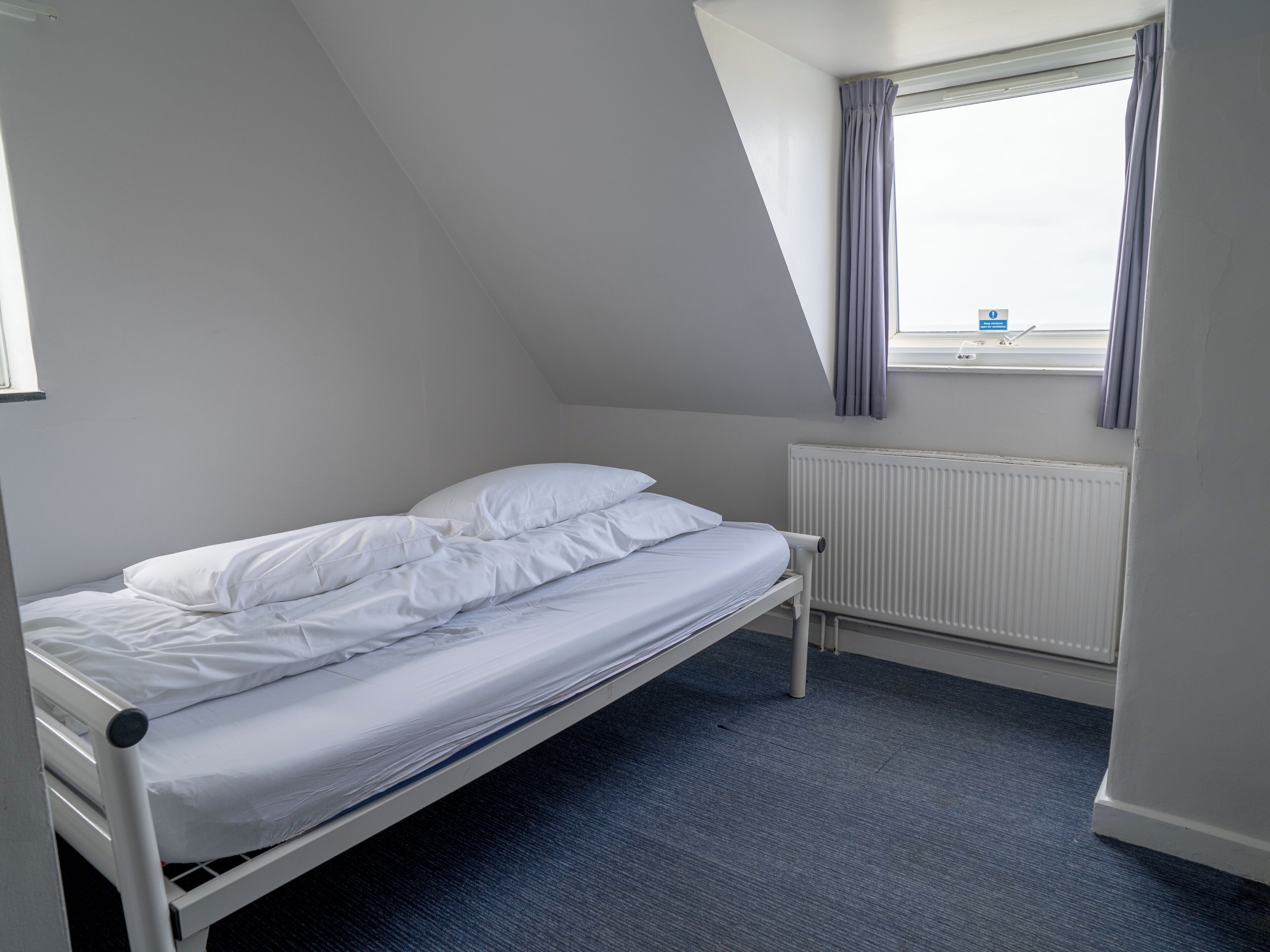 Una cama individual en una habitación con techo inclinado y una ventana con cortinas.