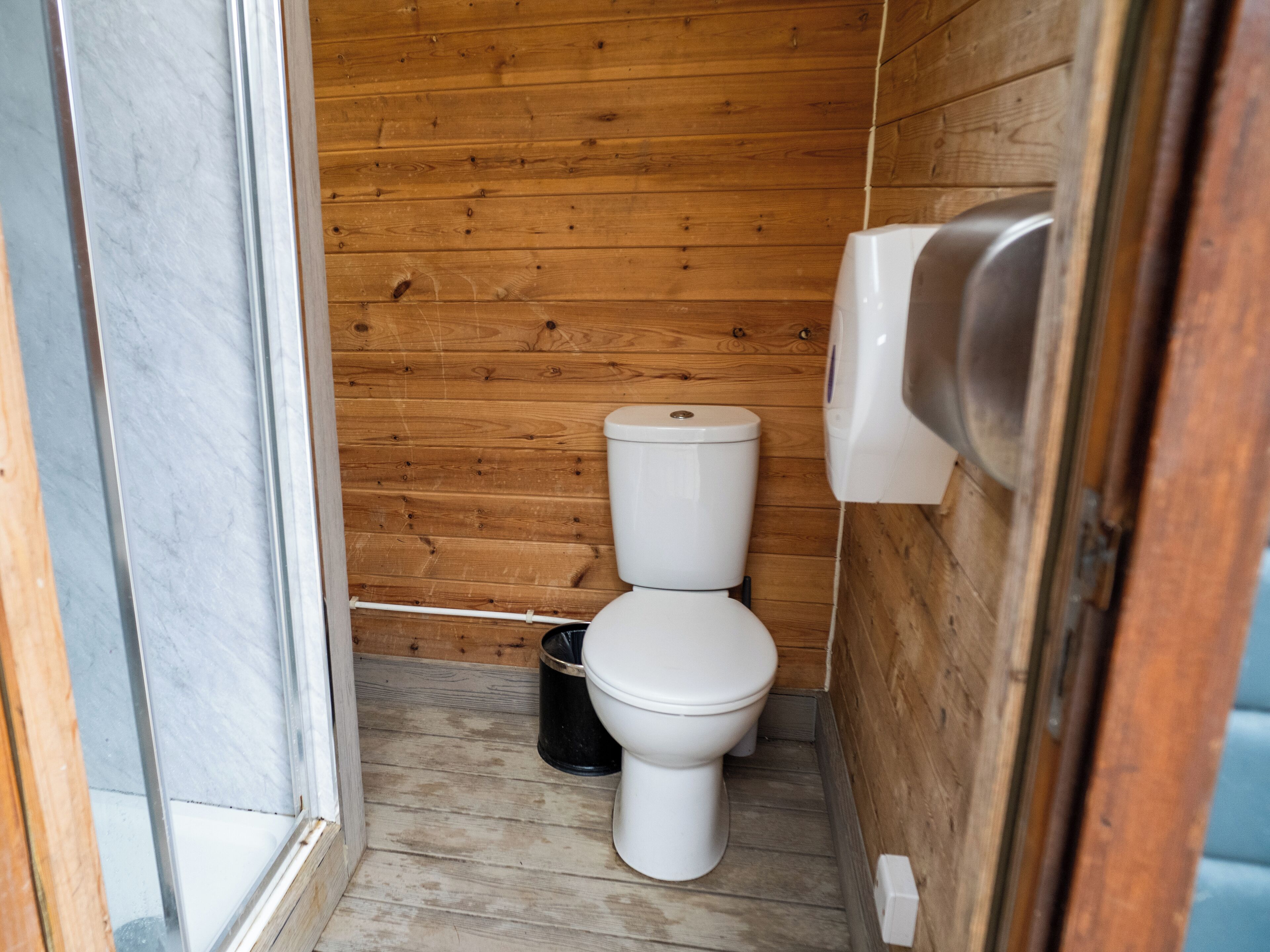 A compact wooden bathroom with a white toilet, a black trash bin, and a hand dryer.