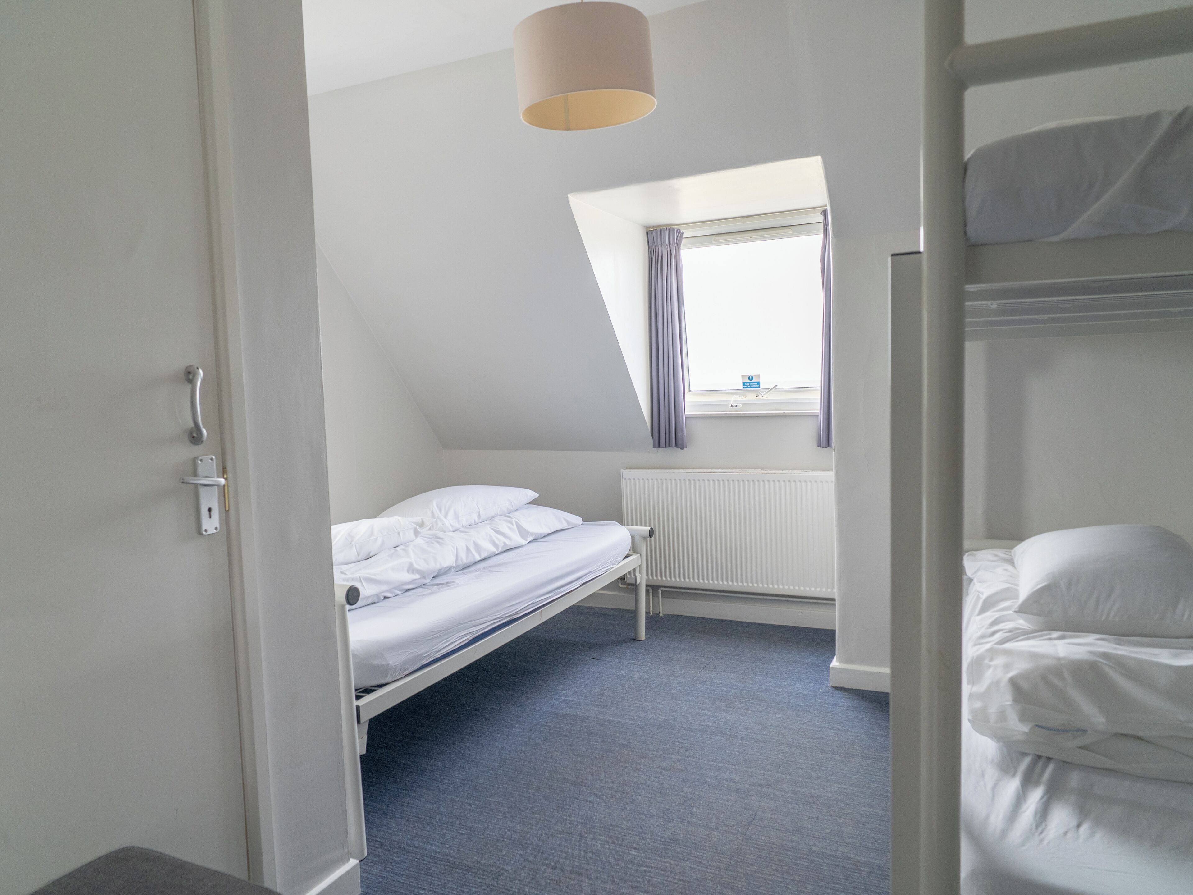 Une petite pièce propre, avec un lit superposé, un lit simple, une fenêtre avec des rideaux, un radiateur et une porte dotée d’une poignée.