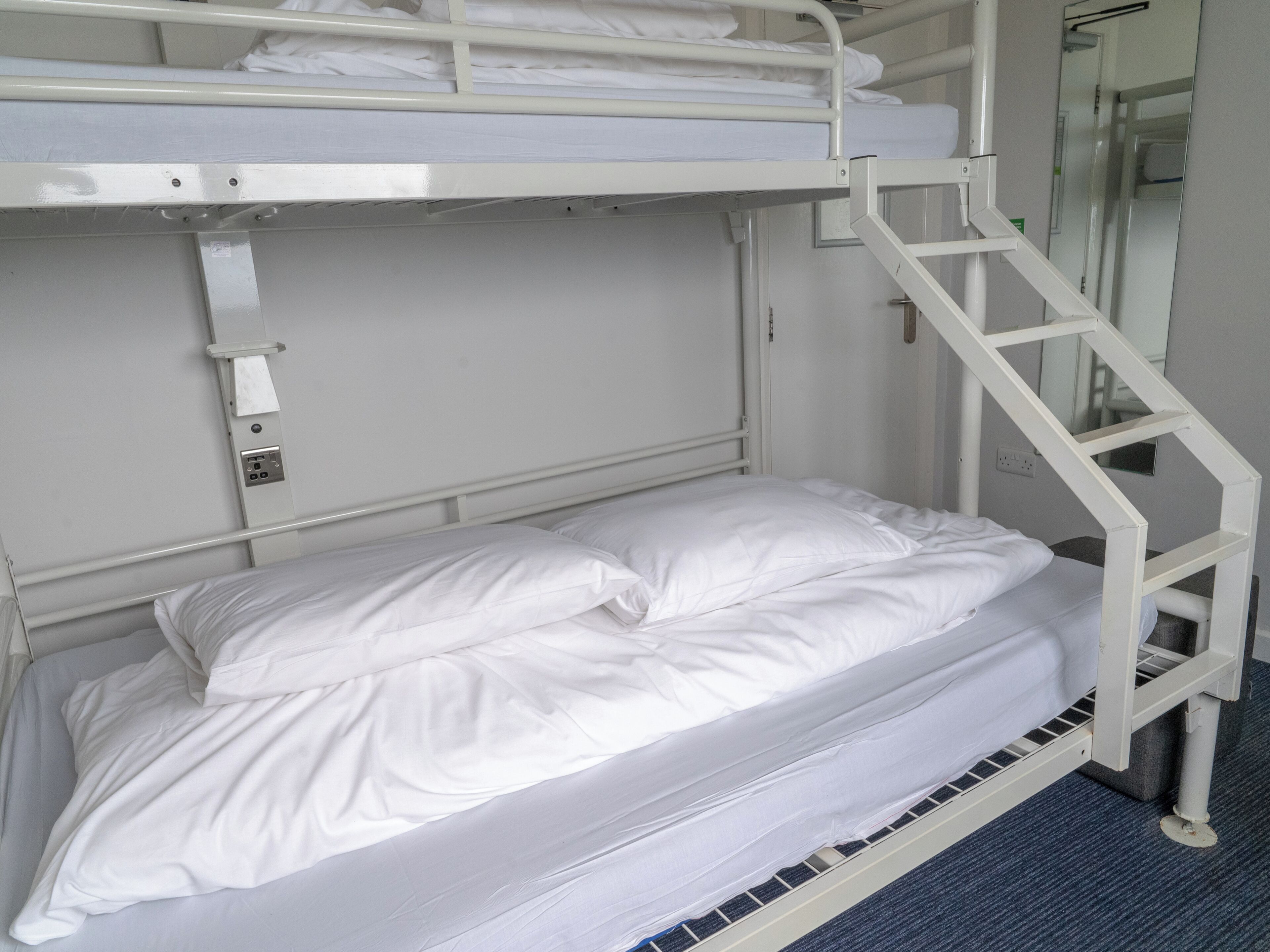 Una cama litera con ropa de cama blanca, una escalera metálica y un enchufe en la pared.