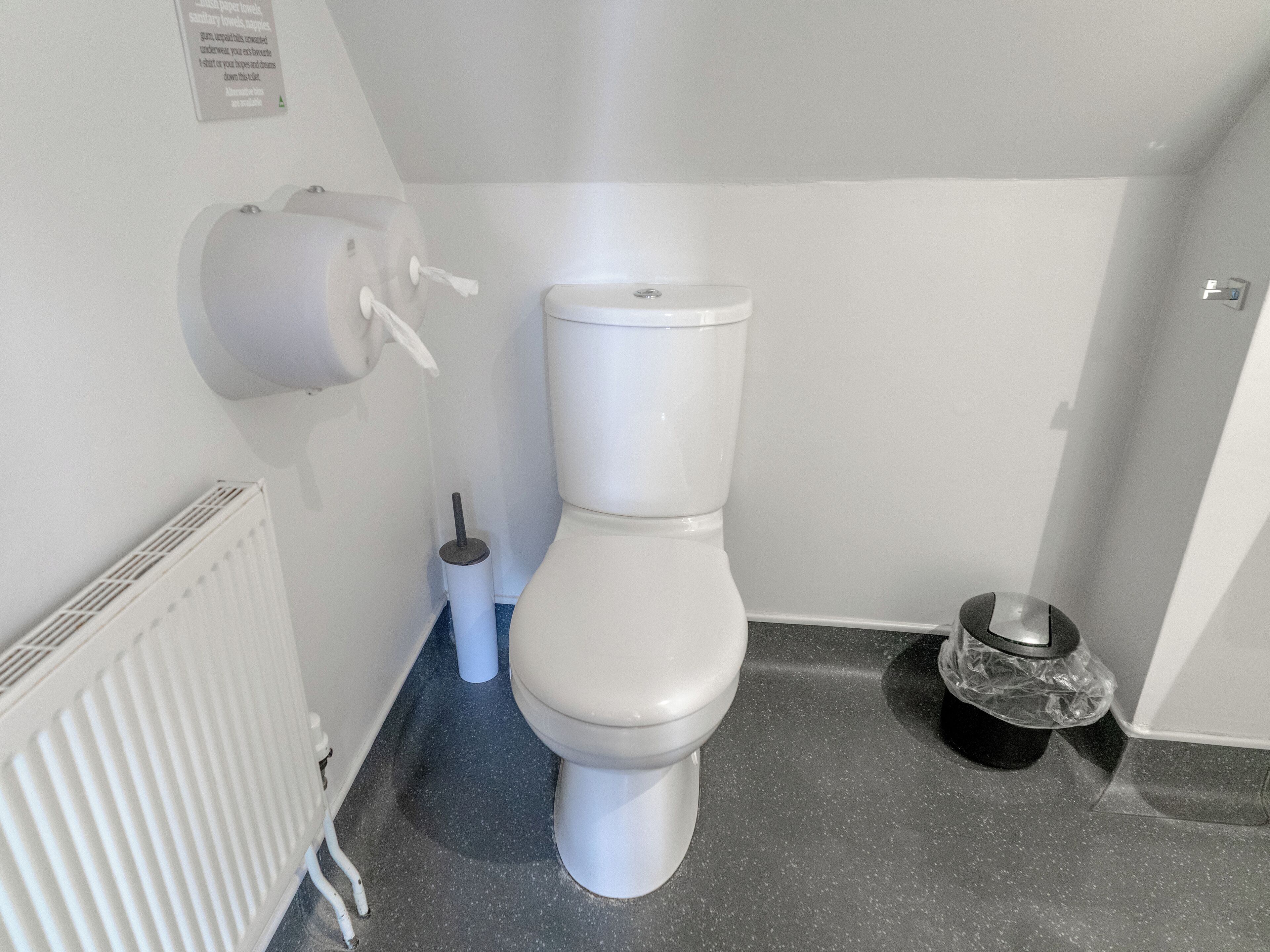 Un baño con inodoro blanco, dispensador de toallas de papel, escobilla de baño y cubo de basura negro.