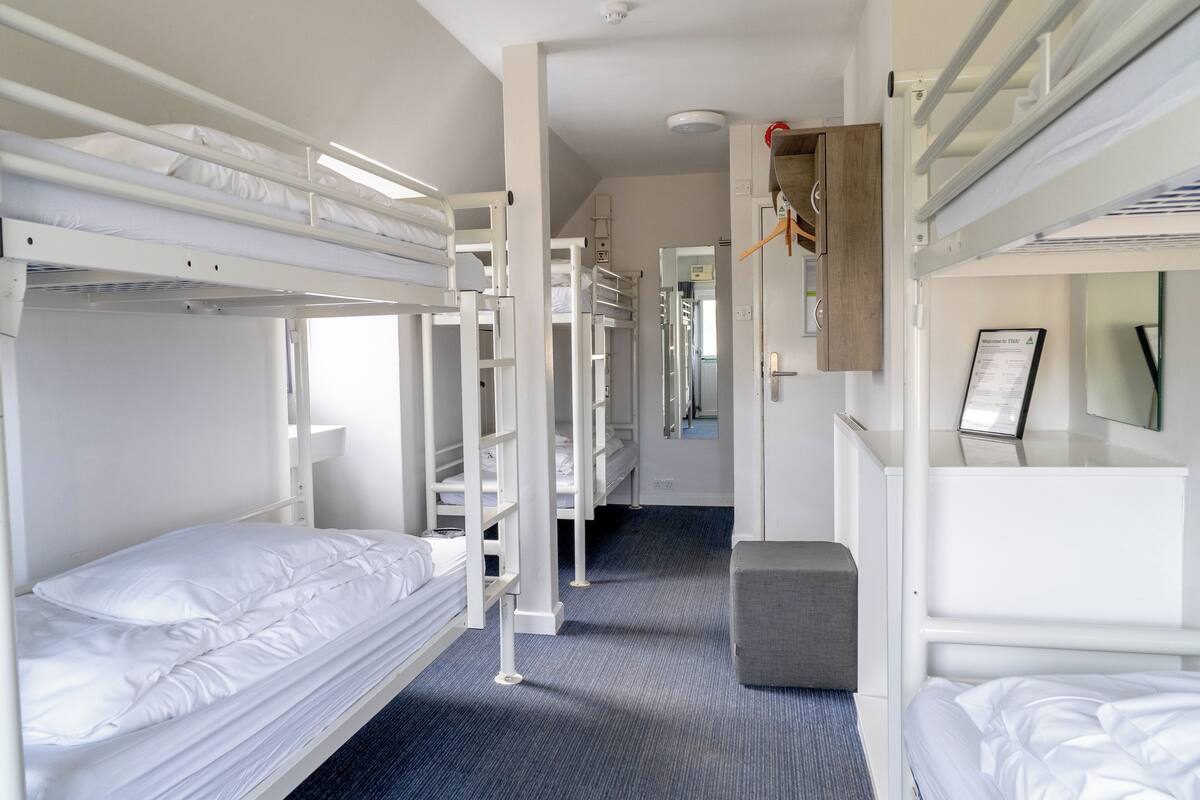 A hotel room with bunk beds, a small gray ottoman, a mirror, and a door leading to a hallway.