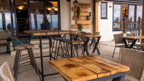 Un área de estar al aire libre con mesas de madera y sillas de mimbre, una escalera que conduce a un nivel superior y una pared decorada con tablones de madera y follaje.