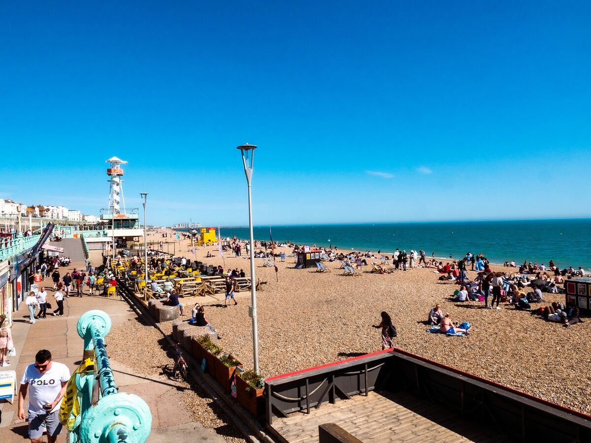 A beach with people sunbathing and walking, a lighthouse, and a boardwalk with shops.