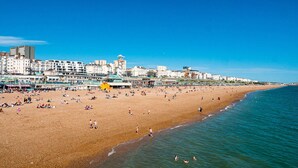 A wide beach with people sunbathing and swimming, a clear blue sky, and buildings in the background. - a&o Brighton Beach (Brighton)