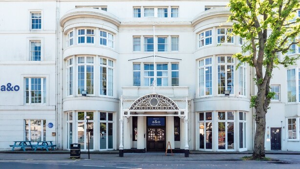A white building with a curved facade, large windows, and a central entrance with a decorative arch. - a&o Brighton Beach (Brighton)
