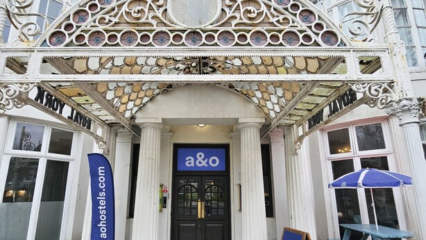 A building with ornate architectural details, a prominent entrance with a sign reading "a&o", and a blue banner advertising "aohostels.com". - a&o Brighton Beach (Brighton)