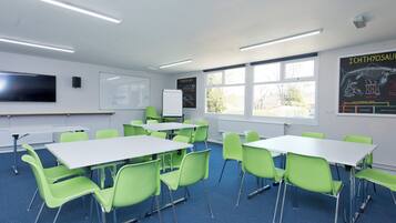 A classroom with green chairs, white tables, a whiteboard, a projector screen, and educational posters on the walls.
