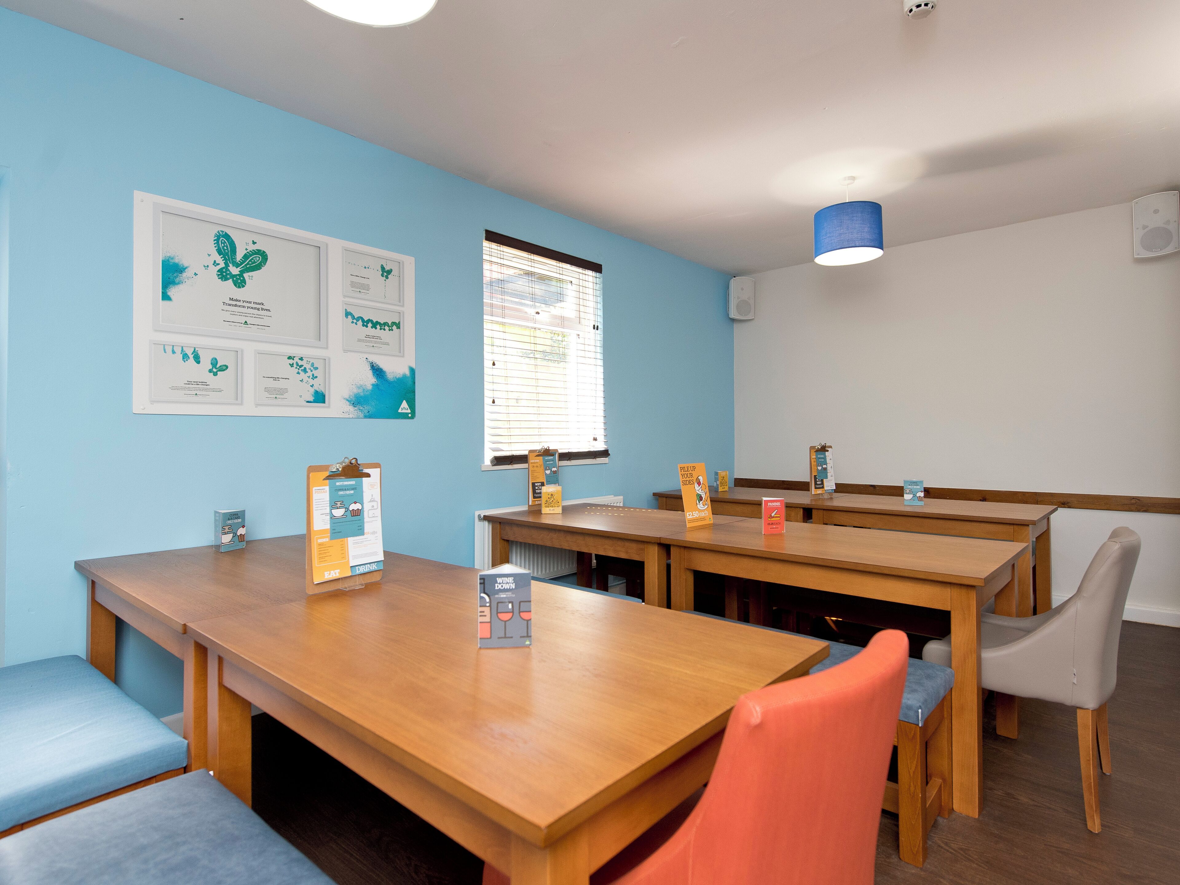 A dining area with wooden tables and chairs, a bulletin board with notices, and a window with blinds.