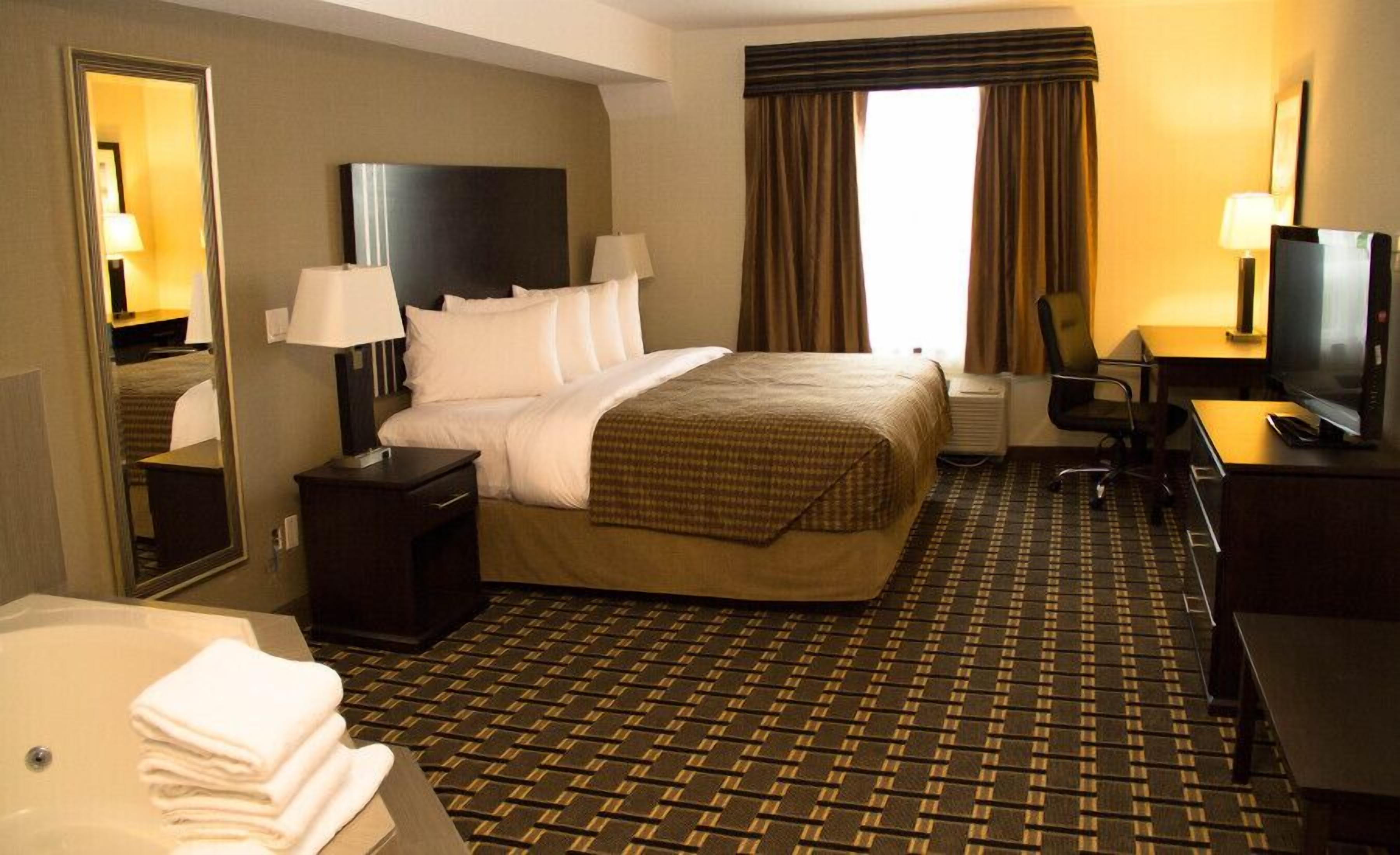 Habitación de hotel con cama, escritorio, televisión, silla, espejo y baño con bañera y toallas.