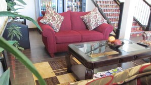Un sofá rojo con cojines estampados, una mesa de centro con superficie de vidrio y una alfombra a cuadros en un salón con una escalera al fondo.